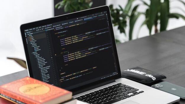 Coderen voor efficiëntere bedrijfsprocessen via automatisering Laptop met code op het scherm voor het automatiseren van bedrijfsprocessen, op een bureau met werkmaterialen.