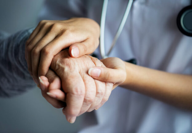 Zorgverlener houdt ondersteunend de hand vast van oudere persoon, symbool voor vertrouwen in langdurige zorg thuis.